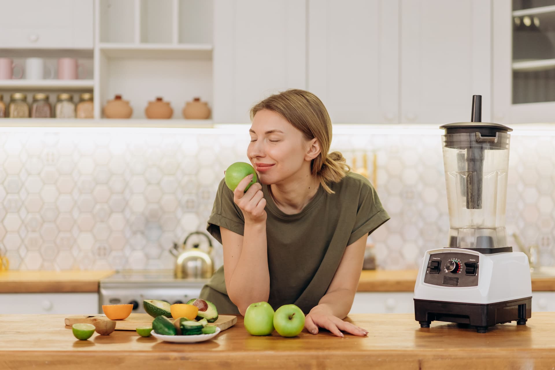 Caucasian woman savoring a green apple in a modern kitchen with fruits and blender.