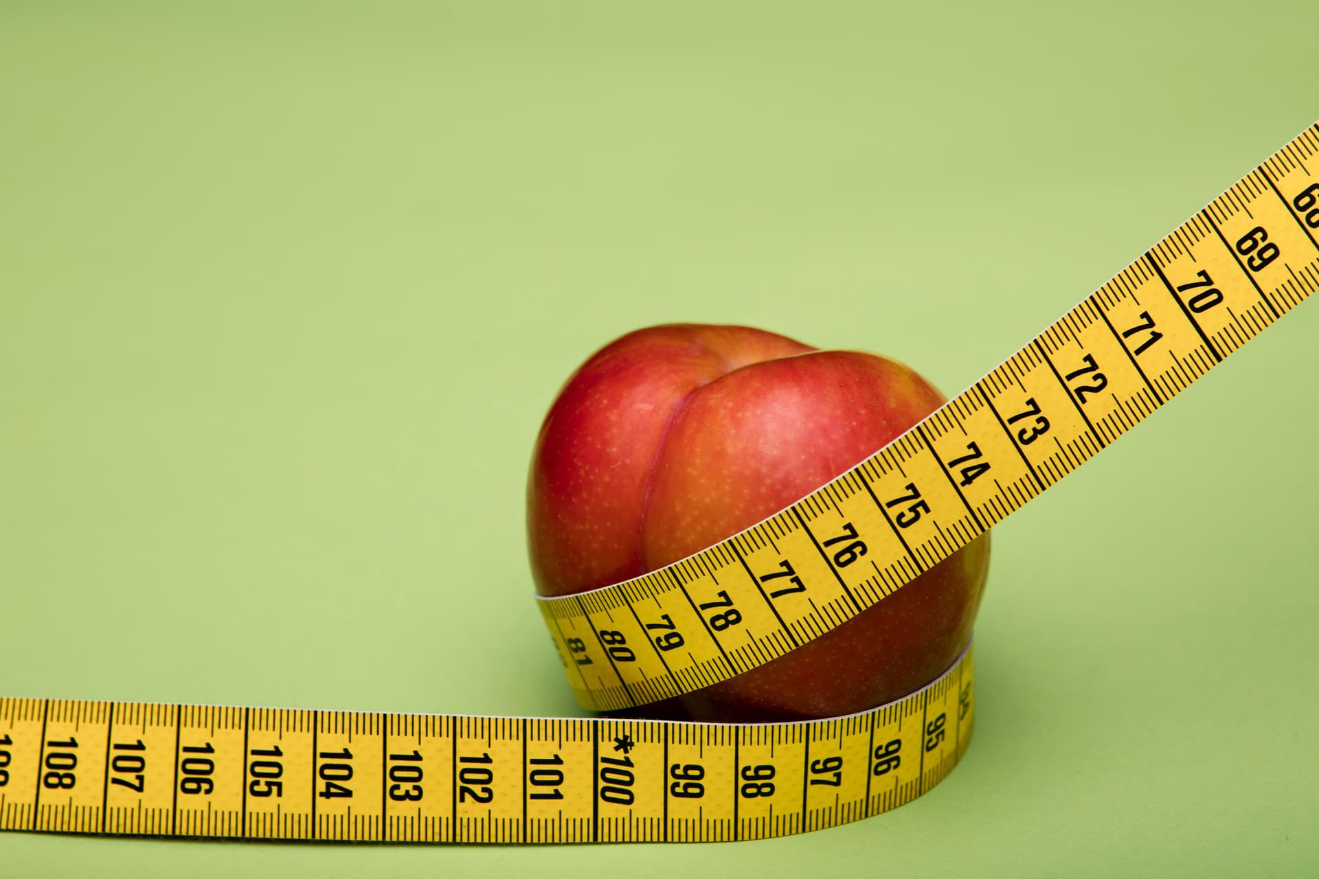 Red apple wrapped in a yellow measuring tape against a green backdrop emphasizes freshness and health.