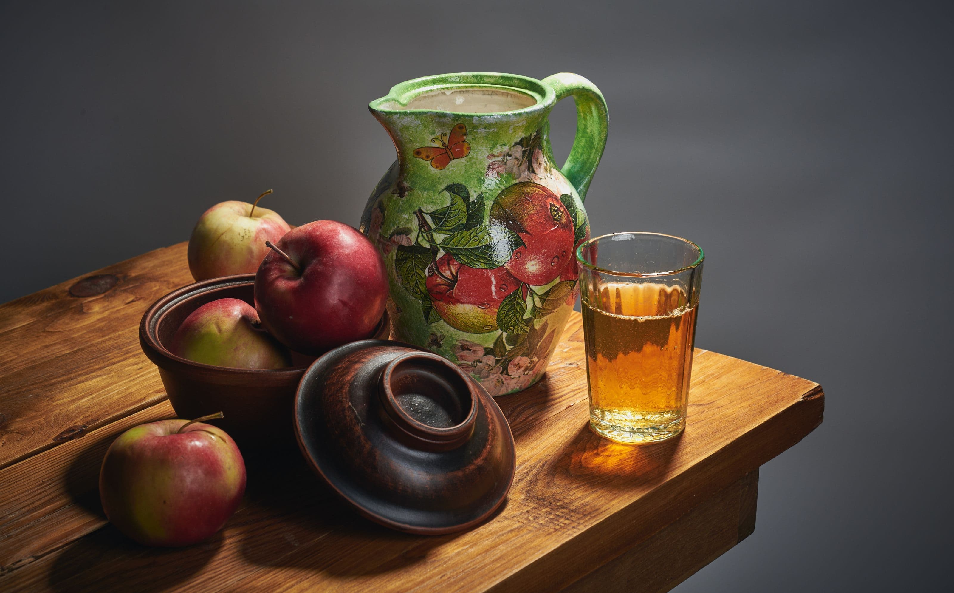 A colorful jug and fresh apples arranged on a wooden table in a classic still life setup.